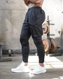 Mann in schwarzer Jogginghose mit Seitentaschen, weiße Sneaker, Fitnessstudio-Hintergrund.