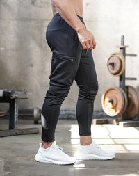Mann in schwarzer Sporthose und weißen Turnschuhen im Fitnessstudio, Hanteln im Hintergrund.
