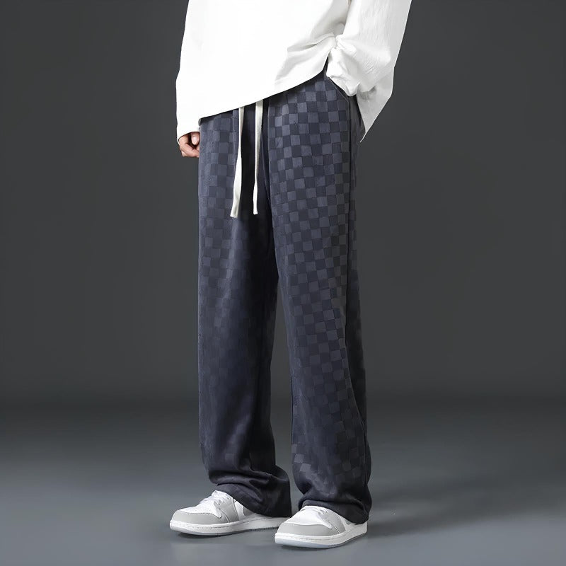 Mann in karierten, dunkelgrauen Jogginghosen und weißen Sneakers vor grauem Hintergrund.