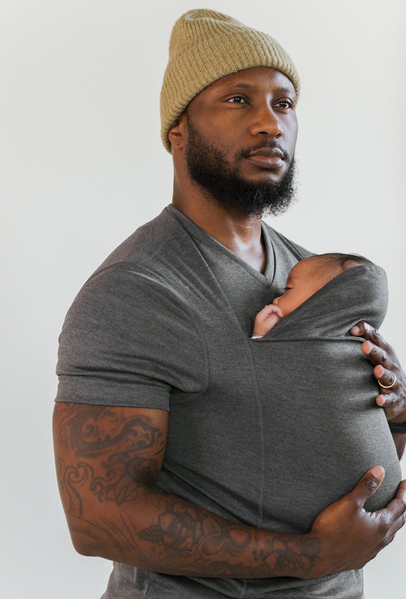 Mann mit grauem T-Shirt und Baby in Tragetuch, Mütze, tätowierter Arm, Familienmode.