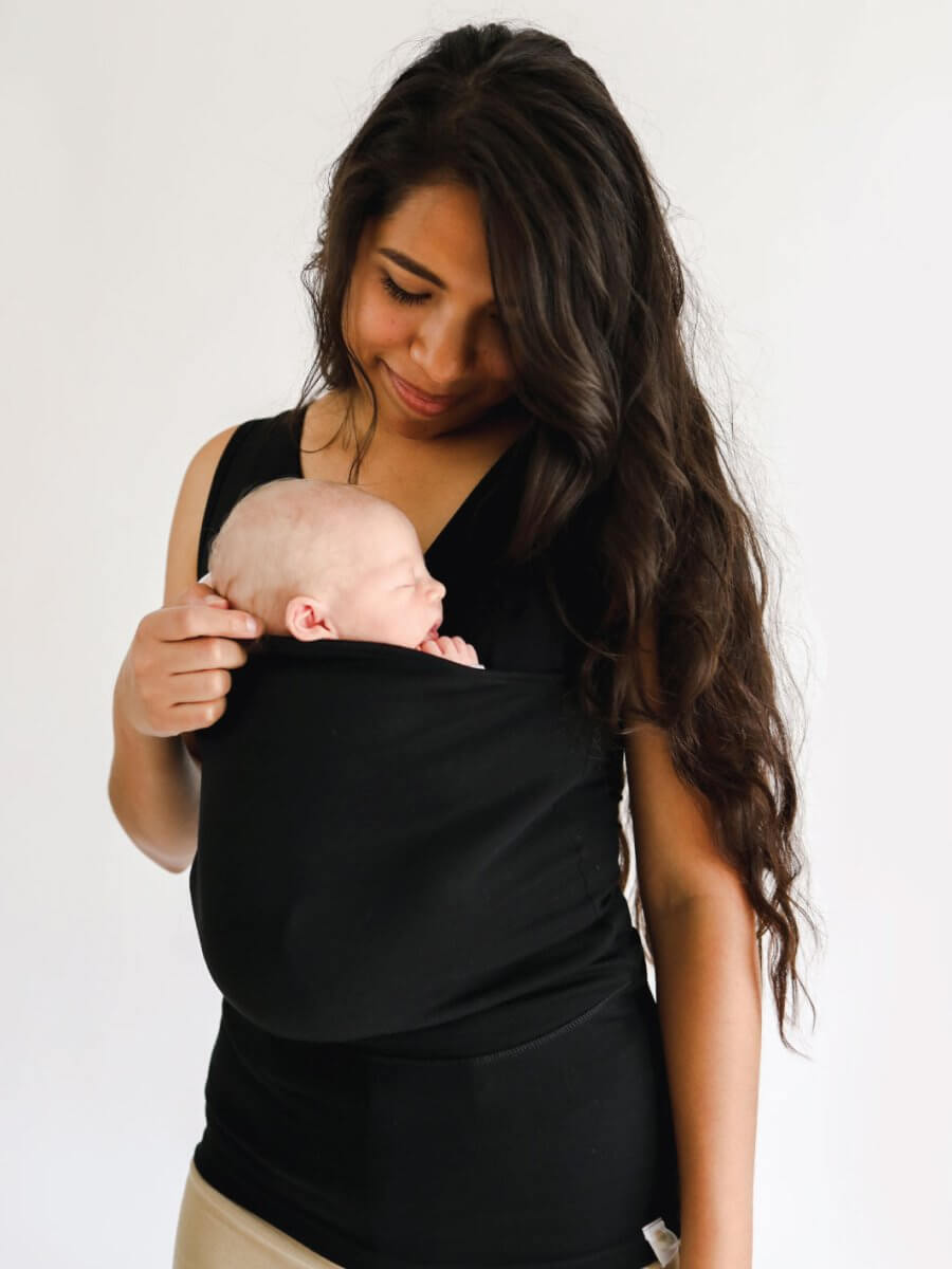 Frau mit schwarzem Babytrage-Shirt, hält Neugeborenes, lächelt, Mutter-Kind-Bindung.